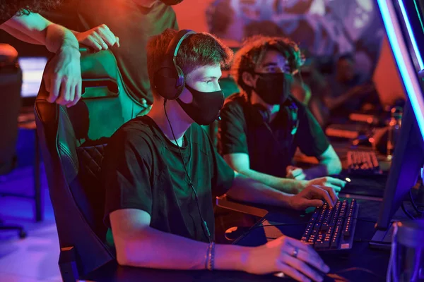 Young man in cloth mask and headphones using computer while participating with professional team in tournament