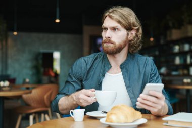 Kahve masasında oturmuş, akıllı telefondan posta okurken kahve içen sakallı yakışıklı bir adam.