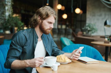 Kafedeki akıllı telefonları kontrol ederken günlük gömlekli, kahve içen ve kruvasan yiyen konsantre genç adam.