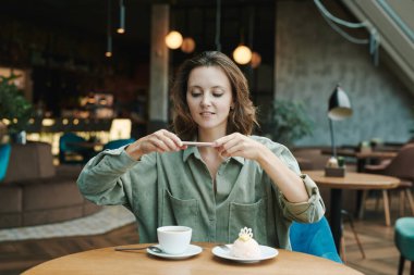 Genç sakallı yönetici kahve fincanı ve kruvasanla masada oturuyor ve kafede akıllı telefon kullanarak iş arkadaşına sesli mesaj kaydediyor.