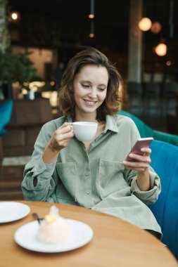 Gündelik gömlekli, rahat bir genç adam modern kafede telefonla konuşurken koltuğunda oturuyor ve başka tarafa bakıyor.