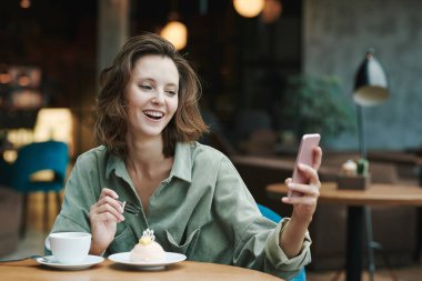 Kafedeki akıllı telefonları kontrol ederken günlük gömlekli, kahve içen ve kruvasan yiyen konsantre genç adam.