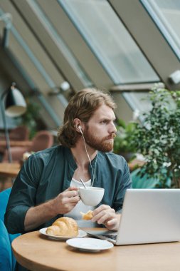 Gündelik gömlekli, rahat bir genç adam modern kafede telefonla konuşurken koltuğunda oturuyor ve başka tarafa bakıyor.