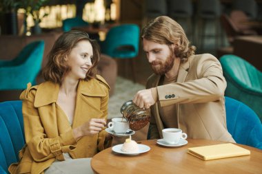 Masada oturan ve kafede onu beklerken iş arkadaşını telefonla arayan sakallı yakışıklı bir genç.
