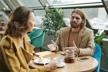 Gündelik gömlekli, rahat bir genç adam modern kafede telefonla konuşurken koltuğunda oturuyor ve başka tarafa bakıyor.
