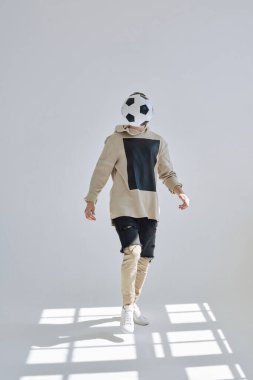 Sokak tarzı giyinmiş tanınmayan bir adam parlak güneş ışığıyla aydınlatılmış beyaz spor salonunda futbol topunu tekmeliyor.