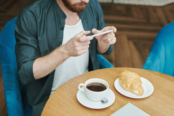 Portrait of serious modern guy with beard sitting at round table in loft cafe and calling by mobile phone
