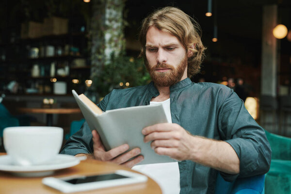 Серьезный красивый парень с бородой сидит за столом в кафе и пьет кофе, читая почту на смартфоне