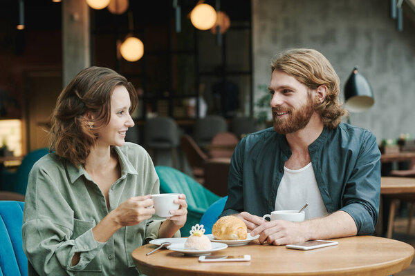Young bearded manager sitting at table with coffee cup and croissant and recording voice message for colleague using smartphone in cafe