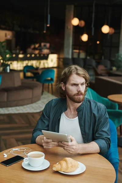 Serious handsome guy with beard sitting at table in cafe and drinking coffee while reading mail on smartphone