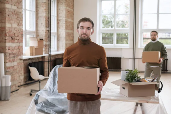 Portrait of serious young bearded man bringing box of office stuff in new office while their company changing workspace