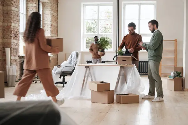 Group of men standing at desk covered with polyethylene and unpacking stuff while their colleagues carrying stuff into new office