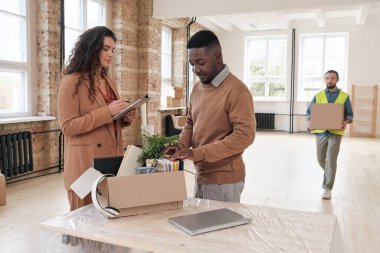 Genç bir kadın eşyalar listesini kontrol ediyor ve siyah bir adam kutulardaki not defterlerini sayıyor. Taşındıktan sonra ofiste eşyaları boşaltıyorlar.