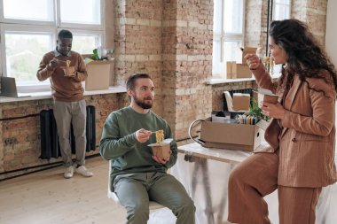 Sandalyeye oturup erişte yiyen genç sakallı adam yeni ofisteki kutularla dolu meslektaşıyla konuşuyor.