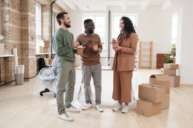 Bir grup gülümseyen, etnik çeşitlilikte genç meslektaşlar yeni ofiste kutuları taşıyorlar ve birlikte Çin yemeği yiyorlar.