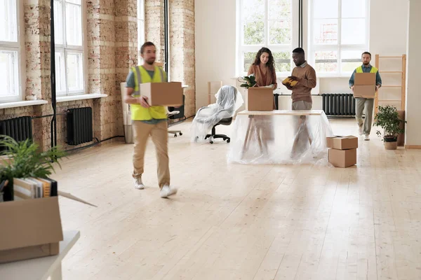 Loaders in vests carrying moving boxes in office while multi-ethnic employees setting up new space