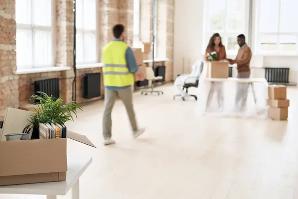 Modern spacious office space with moving stuff and employees unpacking boxes in background, focus on cardboard box with sketchpads