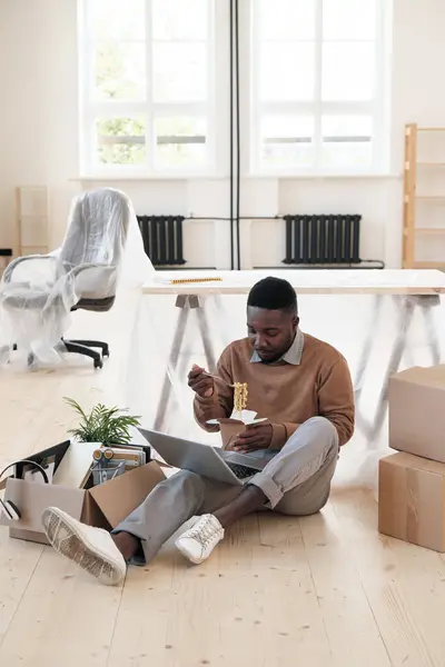 Young Black man in sweater sitting on floor with cardboard boxes and eating noodles while working with laptop in new office
