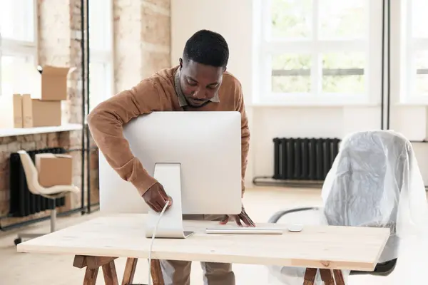 Young Black employee in sweater standing at desk and powering up computer in new office after renovation