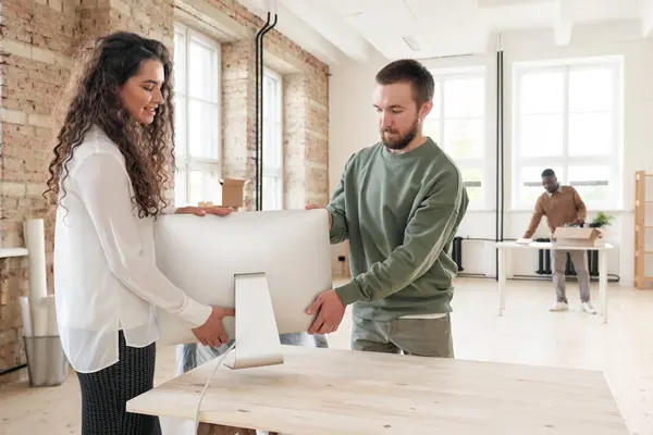 Young bearded man in sweater assisting female colleague to put computer on table in new office after relocation