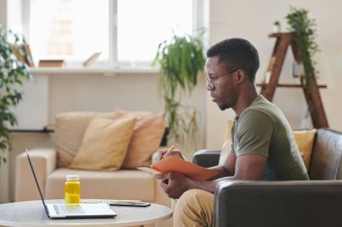 Oturma odasındaki dizüstü bilgisayarın önünde oturan genç siyahi bir adamın doktorla online görüşme yaparken yan görüntüsü.