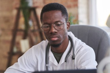 Beyaz önlüklü ve gözlüklü ciddi bir yetişkin doktorun orta boy portresi. Çatı katında oturmuş bilgisayar ekranına bakıyor.