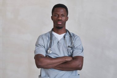 Kollarını kavuşturup kameraya bakarken gri üniforma giyen yakışıklı bir doktorun orta boy portresi.