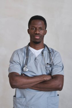 Dikey orta boy, yakışıklı siyahi doktor gri üniformalı, kolları çaprazlanmış kameraya bakıyor.