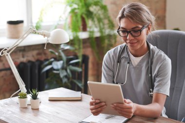 Genç, yetişkin, gözlüklü bir kadın doktor elinde dijital tabletle masada oturmuş video çağrısını dinliyor.