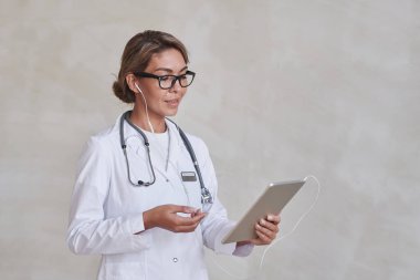 Çevrimiçi randevu için dijital tablet kullanarak gözlük ve kulaklık takan modern kadın doktorun yatay orta boy portresi