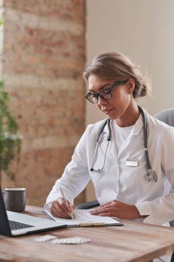 Beyaz önlüklü ve gözlüklü çekici bir kadın doktor çatı katında masa başında oturmuş kağıtlara bir şeyler yazıyor.