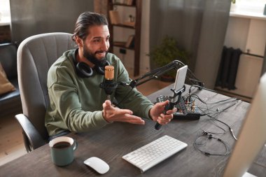 Yatay yüksek açılı orta açılı modern Orta Doğulu sakallı bir adam masasında oturmuş akıllı telefon kamerasıyla video çekiyor.