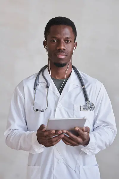 Vertical medium portrait shot of handsome young adult doctor wearing earphones holding digital tablet looking at camera