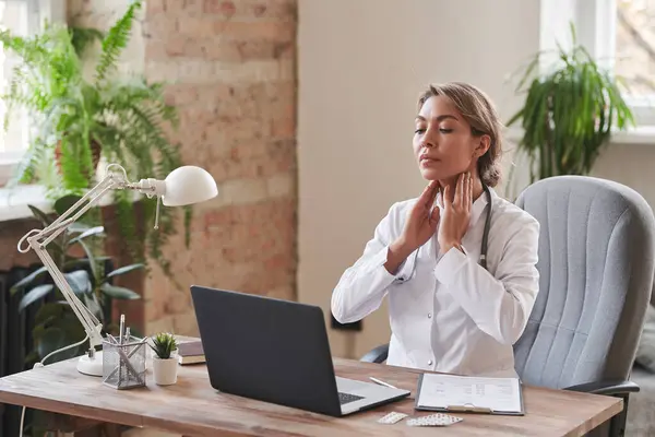 Modern female doctor sitting at desk in office demonstrating how to palpate neck lymph nodes on camera during online consultation