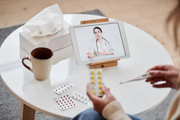 High angle shot of unrecognizable woman having online appointment with doctor using digital tablet holding pills and thermometer