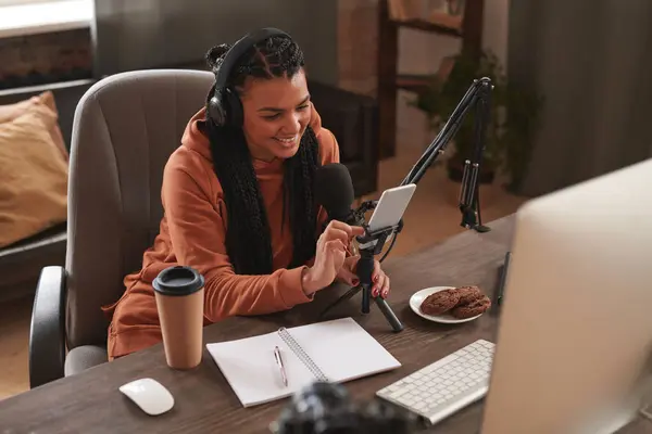 Horizontal high angle shot of happy young female blogger with afro braid hairstyle wearing headphones sitting at desk watching something on her smartphone
