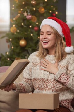 Orta boy beyaz kadın portresi. Günlük sıcak süveter ve kırmızı Noel Baba şapkası giymiş. Kanepede oturmuş Noel hediyesi aldıktan sonra heyecanlanıyor.