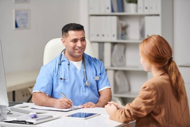 Mavi üniformalı orta yaşlı bir doktor ofisinde oturmuş, tanınmayan uzun saçlı genç bir bayana danışıyor.