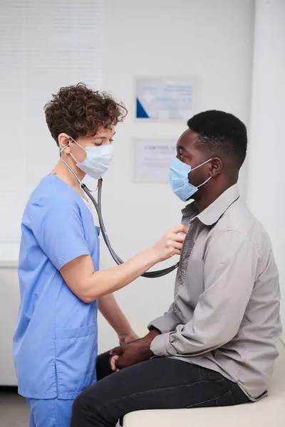 Vertical medium long side view portrait shot of professional female doctor wearing mask examining African American patient using stethoscope