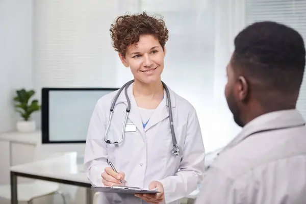 Horizontal over-the-shoulder shot of friendly female doctor wearing white coat listening to patients health complaints and making notes
