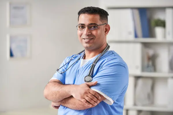 Horizontal medium portrait shot of successful young adult Middle-Eastern doctor wearing blue uniform and eyeglasses standing with arms crossed smiling at camera