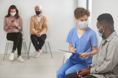 Tanımlanamayan, maske takan, koridordaki koltukta oturan, hastaya sorular soran ve notlar alan bir doktor resmi.