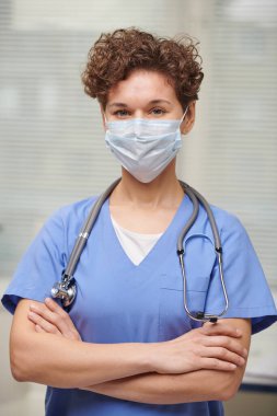 Yüzüne maske takmış, kolları çaprazlanmış kameraya bakarken tanınmayan beyaz bir doktorun dikey belden yukarı portresi.