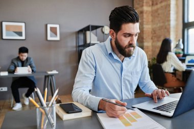 Kafkasyalı bir adamın masada oturup dizüstü bilgisayar kullanarak finans stratejisi üzerinde çalışırken çekilmiş yatay görüntüsü.