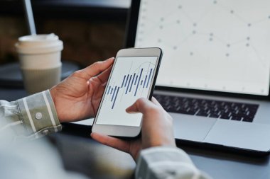 Tanımlanamayan bir kadın dizüstü bilgisayarının önünde oturmuş elinde akıllı bir telefonla hisse senedi alım satım istatistikleriyle çalışıyor.