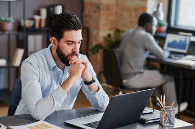 Ofiste çalışan beyaz bir adamın bilgisayar ekranına bakıp bir şey düşünürken orta yatay görüntüsü.