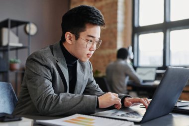 Genç Asyalı gözlüklü bir adamın çatı katında dizüstü bilgisayarla çalışırken çekilmiş yatay görüntüsü.