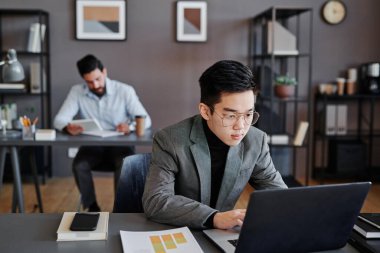 Genç Asyalı gözlüklü bir adamın ofiste dizüstü bilgisayarın önünde oturmuş verilerle çalışırken çekilen yatay görüntüsü.