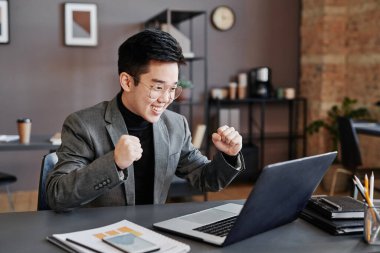 Genç Asyalı komisyoncunun çatı katında masa başında oturup dizüstü bilgisayara bakarken çekilmiş yatay bir fotoğrafı.