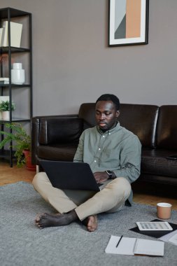 Genç siyahi adamın oturma odasında oturmuş dizüstü bilgisayarla finans projesinde çalışırken dikey çekimi.